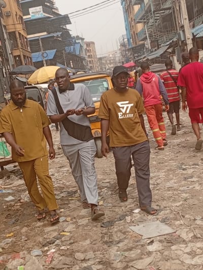 The image depicts a bustling street scene in what appears to be a dense urban environment, likely a market or commercial district in a developing country. The setting suggests a somewhat informal and well-worn area.
In the foreground, three men are walking towards the viewer, slightly angled to the right.
The man on the left wears a mustard yellow or light brown short-sleeved collared shirt and matching trousers, paired with dark brown sandals. He has a dark beard and appears to have a serious or neutral expression.
The man in the center is balding and wears a grey polo shirt with a black panel on one side and a short zipper at the collar. He has light grey trousers with a subtle white stripe along the side seam and dark sandals. He is actively speaking or gesturing with both hands open, his expression engaged.
The man on the right wears a dark brown T-shirt with a prominent white logo that reads "ST ELLESEE." He also wears a dark baseball cap, dark trousers, and dark sandals. His expression is neutral as he looks slightly past the camera.
In the mid-ground, a bright yellow vehicle, possibly a taxi or a small commercial van, is partially visible behind the three foreground men. The letters "TVS" can be faintly discerned on its front. A yellow market umbrella is also visible in the distance to the left of the yellow vehicle.
Several other people, mostly men, are visible in the background, walking in various directions. One man wears a red hoodie and orange or reddish-brown work pants, another wears a red and white striped shirt, and a third is in a plain red T-shirt.
The ground is unpaved and heavily littered with various types of refuse, including plastic bags, paper, and fabric scraps, suggesting a lack of proper waste management or a highly trafficked area. The surrounding buildings are multi-story and densely packed, with visible signs of wear and tear, exposed metal scaffolding, corrugated metal roofs or awnings, and numerous wires and pipes running along their exteriors. The sky appears overcast or hazy, diffusing the light and preventing harsh shadows, which could indicate an early morning, late afternoon, or simply a cloudy/dusty day.
The overall atmosphere is one of daily activity in a busy, working-class urban setting.