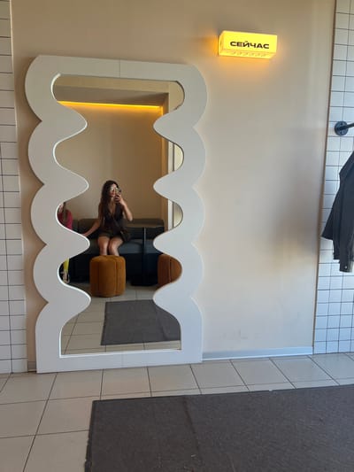 In a well-lit, modern interior space, a woman is captured in a reflection within a uniquely shaped, wavy mirror. The mirror, with its white frame resembling teeth or clouds, is centrally placed against a cream-colored wall adorned with a yellow, illuminated sign that reads "СЕЙЧАС" (which translates to "NOW" in Russian). The sign has decorative cursive elements to its right.

The woman, wearing sunglasses and a dark dress, is seated on a plush, rust-colored ottoman. She is holding a phone to her ear, suggesting she is on a call, and looking into the mirror. Her reflection also shows part of a grey sofa and a dark table behind her. Another person, wearing a pink top and yellow bottoms, is visible in the reflection, sitting on the sofa.

The floor is tiled with light beige squares, and to the right, a dark grey mat is visible. The overall ambiance suggests a contemporary and relaxed setting, possibly a cafe or a lounge area. The lighting, particularly from the "СЕЙЧАС" sign, casts a warm glow on the wall, creating an inviting atmosphere. The time of day is not explicitly indicated, but the bright interior lighting suggests it could be daytime. The scene appears to be indoors, likely in Novoselye, Russia, given the text on the sign and the provided location context.