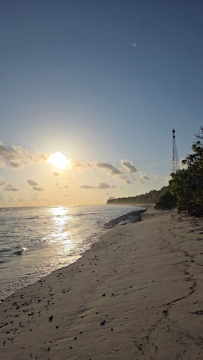 The image depicts a scenic beach in Fuvahmulah, Maldives. The scene is a tropical shoreline with gentle waves lapping the sandy beach, which is speckled with small pebbles. The sun is low in the sky, casting a warm, golden light across the water and the clouds, suggesting it is either sunrise or sunset. A dense line of lush green trees and vegetation borders the beach, hinting at a tropical island setting. In the distance, a tall, metallic tower structure is visible, possibly a communication tower or a landmark. The sky is a clear, pale blue, with a few wispy clouds. The overall mood of the image is tranquil and serene, evoking a sense of natural beauty and peacefulness. There are no people visible in the image. The time of day is likely late afternoon or early morning, given the angle and color of the sun. The weather appears clear and pleasant.