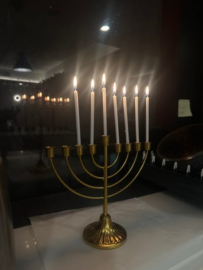 The scene depicts a gold-colored, eight-branched candelabrum with eight lit white candles, placed on a light-colored surface, likely a countertop, within a room in Melbourne, Australia.

The candelabrum is positioned centrally in the foreground. It features a flared, ribbed base and a central stem from which eight curved arms extend to support individual candle holders. Each of the eight holders contains a tall, slender white candle, all actively burning with visible, steady flames.

The candelabrum rests on a light-colored, possibly white, surface that appears to be covered with a slightly textured, light-colored material. The background is a dark, highly reflective surface, likely a glass appliance door (e.g., an oven) or a dark wall. This surface prominently displays reflections of the lit candles as an inverted row of blurry orange-yellow lights. A distinct, bright, circular light fixture is reflected in the upper left of the dark background, with fainter, streaking light reflections extending from the candles. Further to the left, a more diffuse, rectangular light reflection is visible. To the right of the candelabrum, a portion of a dark kitchen appliance or a dish rack is visible, featuring a circular, textured, possibly metallic lid or cover, and a row of small, white, conical items. A light-colored sticky note is adhered to the dark background surface in the upper right section of the frame. No individuals are visible in the image.