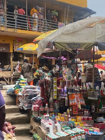 The image captures a vibrant and densely packed outdoor market scene, bustling with activity under bright daylight. The primary orientation appears to be portrait, with a multi-story building on the left and numerous market stalls extending across the center and right.
On the left side of the frame, a yellow-painted building rises, featuring several open balconies or walkways with metal railings. On these upper levels, several individuals are visible: one person sits amidst green bags and what looks like a doll or small child, while others are standing or sitting, observing the scene below or interacting with items. On a lower balcony, a person appears to be handling textiles or clothing. A sign, partially obscured but clearly displaying "POS" in red letters, is attached to the building structure. Towards the bottom of the building, a brown door is visible.
The center and right side of the image are dominated by an intricate network of market stalls. These stalls are covered by makeshift awnings and tarpaulins, with one prominent light-colored (white/khaki) tarpaulin featuring the word "CNGA" in blue lettering. Below it, a yellow banner or sign hangs. The stalls are overflowing with a diverse array of merchandise, creating a cluttered but dynamic visual.
Among the goods displayed are:
* Numerous packaged items on shelves, including detergents, cleaning supplies in white plastic bottles with red caps, various small household items, and toiletries (e.g., toothbrushes, razors).
* Bundles of items hanging from racks, such as long strings of red beaded necklaces or rosaries, colorful sachets or small packets (possibly spices or candies), and plastic bags containing various food items like dried fish or nuts.
* Piles of fabrics or clothing items are stacked in some areas, along with various other small consumer goods.
* Cardboard boxes are utilized for display and storage, adding to the impromptu nature of the market setup.
Several people are interspersed among the stalls. One individual, wearing a patterned garment (likely a local fabric design), is seated behind a stall, seemingly attending to the merchandise. Other figures are partially visible, suggesting a lively environment of vendors and shoppers.
In the very bottom right corner of the frame, a fragment of a person's head and shoulder is visible, clad in purple fabric. Adjacent to this, a large cut of raw meat or produce rests on a surface, indicating the presence of food vendors.
The scene conveys a sense of a busy, possibly congested, commercial hub, typical of a street market in an urban or semi-urban setting, filled with everyday necessities and local goods. The bright lighting suggests it is daytime, and the overall impression is one of vibrant economic activity.