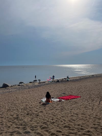 A wide shot captures a beach scene under a bright, hazy sky. In the foreground, a person sits on a towel on the sand, facing away from the camera, dressed in a swimsuit. To their right, a red towel is spread out on the sand. The beach transitions to a rocky shoreline bordering a calm body of water. Several people are visible in the water, some wading and others standing. In the distance, the sun reflects brightly off the water's surface. Several inflatable objects, including a pink flamingo, are present near the water's edge. The location is identified as the city of Luzhki, Russia.