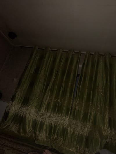 A dimly lit interior scene features a pair of olive-green curtains with a decorative embroidered border. The curtains are hung from a rod near the ceiling and are partially drawn, revealing a dark gap between them. The fabric of the curtains appears to have a subtle shimmer. The wall behind the curtains is textured with a subtle pattern, and a light switch is visible on the wall to the left. The ceiling is dark and appears to be painted a dark color. The floor is covered with a rug that has a visible pattern in dark colors. A dark object, possibly a speaker or light fixture, is attached to the ceiling in the upper left corner of the image. The scene is shot from a low angle, looking upwards. The location is identified as a residential interior in Dushanbe, Tajikistan.