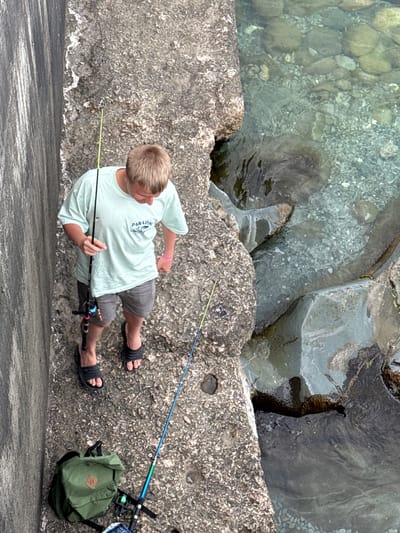 This is a high-angle, full-shot photograph of a young boy fishing on a rocky shore. He is standing on a rough, gray concrete embankment, facing towards the water. The boy has light brown hair and is wearing a light blue t-shirt with blue lettering that appears to say "PARADISE", grey shorts, and black flip-flops. He is holding two fishing rods. One rod is extended in front of him, and the other is held in his right hand. A small green backpack is situated to his right, near another fishing rod that is laid down.
The background shows the clear, shallow water of the sea. The water is calm and reveals a rocky seabed with various sizes of smooth, rounded stones. The rocks on the shore are rough and uneven, with some appearing to be covered in small, light-colored organisms. The lighting suggests it is daytime, likely with sunny conditions given the clear water and shadows. The overall scene depicts a peaceful, outdoor activity of recreational fishing.
The location context is identified as Partenit, Ukraine. There are no strong emotional cues evident, beyond the focused posture of someone engaged in fishing.