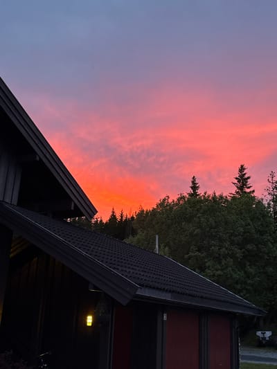 The image depicts a scenic sunset over a wooded area and a dark-colored building. The sky transitions from a pale blue at the top to vibrant shades of pink and orange near the horizon, with wispy clouds scattered throughout. A portion of a dark-colored roof and the side of a building are visible in the foreground on the left. The building appears to be constructed with dark wood siding. A light fixture with a yellow glow hangs from the eaves of the building. Behind the building, a dense forest of green trees forms a dark silhouette against the colorful sky. The image was taken in the city of Åmot, Norway, during twilight. There are no people visible in the image.