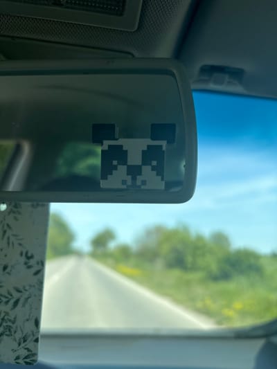 The image captures a view from inside a car, focusing on a sun visor mirror. Affixed to the center of the mirror is a black and white pixelated sticker, depicting a block-art character that resembles a panda. The mirror itself has a dark, possibly plastic, frame.
Through the car's windshield, a bright, sunlit outdoor scene is visible, rendered in a soft blur. A clear, vibrant blue sky occupies the upper portion of the background. To the right, lush green trees and foliage suggest a natural or rural environment. A light-colored road or path can be seen stretching into the distance amidst the greenery. The ambient light indicates it is daytime, likely mid-day or afternoon, under clear weather conditions.
In the lower right foreground, partially visible beneath the mirror, is a white object adorned with a pattern of green leaves, possibly a tissue box or another car accessory. The photograph is taken from a perspective within the vehicle, looking forward and slightly to the passenger side. Given the location context, the scene is set in Trilistnik, Bulgaria. No discernible text is present in the image.