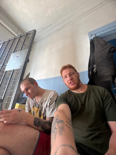 Two men are inside a room with light-colored walls and a white ceiling. On the left, a man with a shaved head and tattoos on his arms wears a t-shirt with a graphic design. He is looking down. To his right, a man with red hair and a beard, also with tattoos, wears a green t-shirt and is looking at the camera. A dark-colored jacket is hanging on a blue door frame behind him. A metal grate structure is visible on the far left of the image. The overall environment appears utilitarian and somewhat worn.
