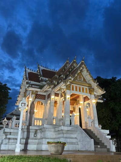 A white, ornate building with tiered roofs and gold accents stands against a dark blue, cloudy sky at dusk. The building, likely a temple or shrine, has intricate carvings and is illuminated by warm interior lights visible through open arches. A set of stone stairs leads to the entrance, and a white lamp post stands in the foreground to the left. The surrounding area is dark, with trees visible on the right side. The scene appears to be located in Tha Khlong Town Municipality, Thailand, given the architectural style. There are no people or animals visible in the image. The ground is wet, suggesting recent rain.
