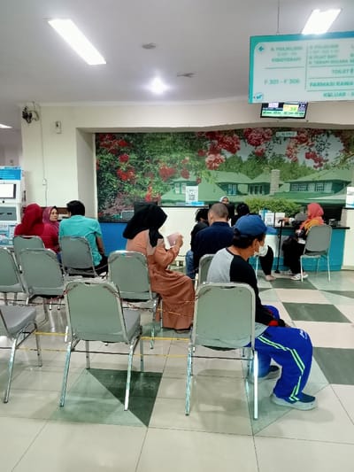 The scene is a waiting area in a hospital, located at coordinates (-6.76, 111.015) in Kecamatan Margorejo, Indonesia. Seven individuals are seated in rows of chairs, facing a reception desk. The room features a tiled floor with a checkered pattern. The wall behind the chairs displays a mural of a lush green garden with red flowers, a white picket fence, and a small white house. A sign above the reception desk indicates that it is a clinic offering physiotherapy services. There is a digital clock displaying the time 09:43:02. A blue sign with white text announces that there are a variety of services offered in the building, including physiotherapy, a pharmacy, and a toilet. There is a metal railing separating the waiting area from the reception desk. The waiting area is brightly lit with fluorescent lights. Some of the individuals seated are wearing face masks, suggesting the scene could be during a pandemic or a period of time when masks were required in public spaces. The subjects in the photo are all of Asian ethnicity. There are no discernible interactions happening between individuals. The scene appears to be an everyday occurrence in the hospital.