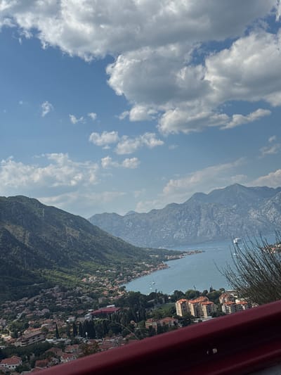 This is a vertically oriented, wide-angle photograph taken from a high vantage point, looking down and across a bay or large body of water towards a mountainous landscape. The sky occupies the upper left portion of the frame and is a bright blue with scattered white clouds. The lower right portion of the frame is dominated by a steep, tree-covered mountainside dotted with numerous buildings, suggesting a town or village clinging to the slopes. These buildings have terracotta roofs and are clustered together, creating a dense settlement. The water in the middle ground appears calm, with several small boats visible on its surface. In the background, a long, jagged mountain range stretches across the horizon, partially obscured by atmospheric haze, giving it a muted blue-grey appearance.
The sun appears to be shining, casting a bright light and creating a clear, albeit hazy, atmosphere. The time of day is likely daytime, possibly late morning or early afternoon, given the brightness of the sky and the clarity of the scene. The overall impression is one of a scenic coastal or lakeside town nestled within a dramatic natural landscape. The photo is framed on the right by a thick, dark red, curved object that seems to be part of a structure or railing, perhaps a balcony or a vehicle. There is no visible text in the image.
