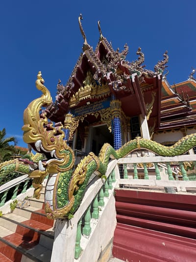The image shows an ornate, multi-tiered roof of a temple in Baan Plai Laem, Thailand, under a clear blue sky. In the foreground, a large, golden and green dragon sculpture wraps around a staircase. The dragon has a prominent head with fangs and appears to be guarding the entrance to the temple. The staircase has red and white checkered steps and is flanked by a green balustrade. The temple itself is richly decorated with intricate carvings and golden accents. The overall scene conveys a sense of grandeur and cultural heritage.