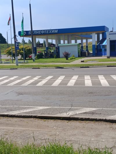 The image depicts a modern gas station with a blue and yellow canopy, identified by the Cyrillic text "БЕЛНЕФТЕХИМ" on the canopy. Two flags are visible, one with the Belarusian national colors and another green flag. The gas station is located in Polatsk, Belarus. In front of the gas station, there is a pedestrian crossing marked with white stripes on the asphalt road. The surrounding area is grassy with some trees and bushes. Several vehicles, including a truck and a car, are parked near the gas station's service area. A person is walking near the service area. The sky is clear and blue, indicating a sunny day.