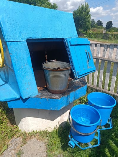 In Zhyhalivka, Ukraine, a rustic outdoor scene unfolds around a weathered blue well. The well's structure, painted a vibrant, albeit chipped, blue, features an open top where a metal bucket hangs by a chain. The bucket appears to be made of galvanized metal and shows signs of rust around its rim. Beside the well, two bright blue plastic buckets are visible. The closest one is partially filled with clear water, its surface rippling and reflecting the sunlight. The second blue bucket, positioned slightly behind and to the right of the first, also contains water. The well and buckets sit on a surface that transitions from a rough stone path to green grass. Behind the well, a weathered wooden fence with white-painted posts runs horizontally, suggesting a property boundary. In the background, lush green trees and a hazy sky with scattered clouds complete the rural setting. The scene depicts a functional, yet aged, water collection point in a village environment.