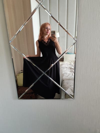 A woman with reddish-blonde hair stands in front of a mirrored wall, taking a selfie with her phone. She is wearing a sleeveless black dress with lace detailing around the neckline and a full skirt. Her right hand is holding her phone, and her left hand is lifting the skirt of her dress. The mirrored wall is composed of diamond-shaped panels. In the reflection, a bed with white and grey linens is visible, along with a light-colored headboard and a window with a white curtain. To the left of the woman in the reflection, a pale yellow bag is on the floor. The room appears to be a hotel room.