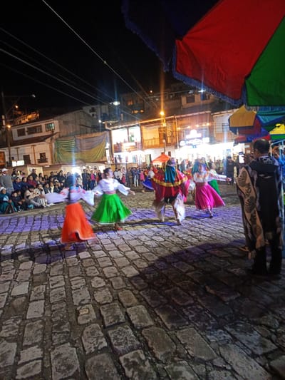At night in what seems to be a town square in El Cisne, Ecuador, a group of people in colorful traditional clothing perform a dance on a cobblestone street. The dancers are wearing skirts in red, green, and pink colors, and one is wearing a peculiar mask and cloak. A crowd of onlookers is seated on the side, and some are filming the performance. A large colorful umbrella is visible on the right side of the frame. Buildings lit by electric lights form a backdrop. The street is lit with overhead lights.