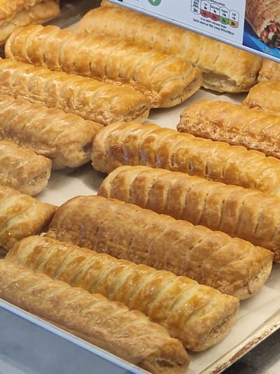 The image shows a tray of freshly baked sausage rolls. They appear to be arranged in neat rows within the tray, their golden-brown crusts and visible pastry layers suggesting a flaky texture. There are several sausage rolls in varying lengths and thicknesses. The tray seems to be made of a light-colored material, possibly cardboard or aluminum. The top left portion of the image contains a partially visible blue and white box with nutritional information. The location is unspecified and there is nothing to indicate the city of Pity Me, United Kingdom.