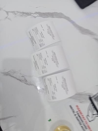 The image shows three receipts laying flat on a white marble table. The receipts appear to be from a business called "EMBASSY HOTEL & SUITES." The top of each receipt is cut off, only the bottom part of the receipts are visible. The image contains no people or other objects. The location is a table in a room at the Embassy Hotel and Suites in Ujevwu, Nigeria, based on the provided coordinates. The environment is an indoor setting. The image appears to be taken from a slightly angled downward position.