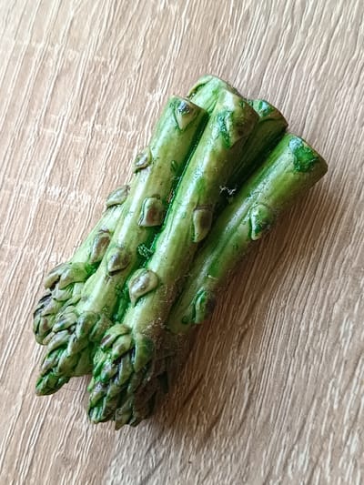 A cluster of five asparagus spears lies diagonally on a light brown wooden surface. The spears are green with small, brown, bud-like structures along their length. The wooden surface has a pronounced wood grain pattern running vertically from top to bottom. The image is a close-up shot, with the asparagus occupying the majority of the frame. There are no people or any other objects visible. The lighting appears to be from above and slightly to the left, casting faint shadows. The scene is located in Tebras, Latvia.