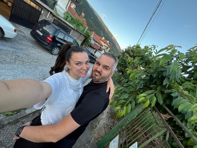 A selfie captures two individuals, a woman and a man, posing on a paved outdoor path in Karlovo, Bulgaria. The woman, with dark hair in a high ponytail and wearing a white t-shirt, smiles at the camera, holding her left arm extended for the shot. Her right arm is around the man. The man, with short grey hair and a beard, wearing a black t-shirt, makes a humorous grimace towards the camera with his right arm around the woman's back. In the mid-ground, a residential street is visible, featuring a parked black Volkswagen van with Bulgarian license plate "PB 8659 PA" and the partial front of a white car. Behind the street, houses with tiled roofs are nestled into a hillside covered with greenery, leading up to a large, forested mountain under a clear blue sky. Overhead power lines are visible against the sky. To the immediate right, a green metal fence and dense green foliage are prominent.