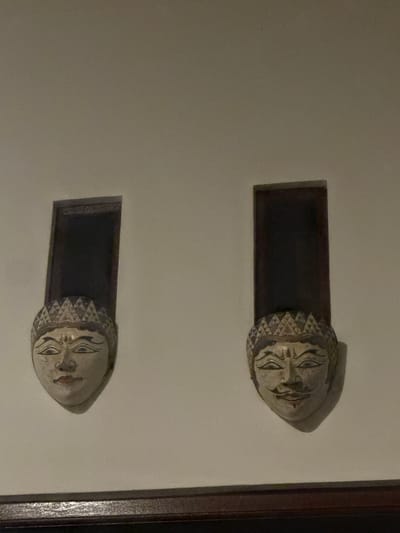 Two carved wooden masks are mounted on a light-colored wall in Abiansemal, Indonesia. Each mask is attached to a dark, rectangular panel extending upwards. The masks depict stylized human faces with painted features. The mask on the left has closed eyes and red lips, while the mask on the right has open eyes, a mustache, and a smiling mouth. Both masks wear decorative headdresses. A dark wooden trim runs along the bottom of the image.