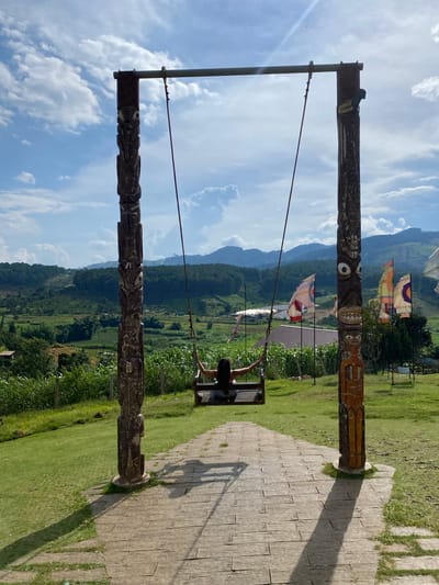 This media file captures a person on a swing with a scenic mountain backdrop. The swing set is constructed with two large, intricately carved wooden poles, each adorned with stylized faces and patterns. A person, with dark hair, is seated on the swing, which is suspended by ropes from a metal bar connecting the tops of the wooden poles.
The background reveals a lush green landscape with rolling mountains fading into the distance. Several colorful flags with geometric patterns are visible, adding a festive touch to the scene. The sky is a vibrant blue with fluffy white clouds, suggesting a pleasant day. The ground near the swing set is paved with rectangular stones, and to the right, a manicured grassy area is visible, casting long shadows from the swing structure. The overall impression is one of outdoor recreation and appreciation of natural beauty. The carved poles and flags hint at a cultural or tourist destination, likely in Vietnam, given the location context. The person on the swing appears to be enjoying the view and the gentle motion. The lighting suggests it is daytime, likely mid-morning or afternoon, with the sun casting distinct shadows. No visible text is present in the image.