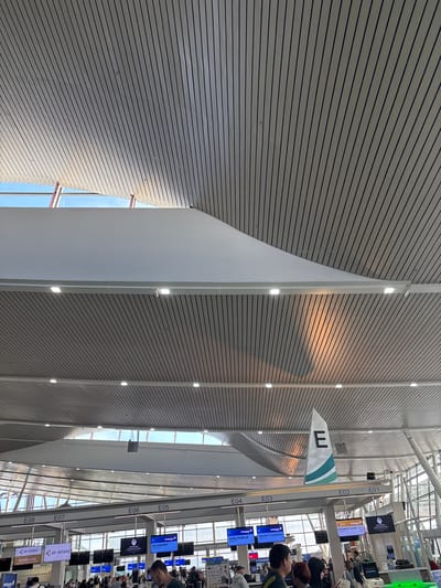 This image captures a bright and modern airport terminal, identified as being in Tambon Sa Khu, Thailand. The dominant feature is a high, intricately designed ceiling composed of light-colored, slatted panels that run horizontally across much of the frame. Integrated strip lighting is visible within these panels, illuminating the space, while large skylights allow natural daylight to stream in.
On the right side of the terminal, a series of check-in counters are clearly visible, sequentially labeled from "E01" to "E08". Above these counters, numerous digital display screens show airline information. Visible text on these screens includes "malaysia airlines" and "air astana" logos, along with service categories such as "BUSINESS CLASS CHECK IN," "PRIORITY COUNTER CHECK IN," "WEB/MOBILE CHECK IN," and "ECONOMY CHECK IN." A partially visible word "PLENGE" also appears on one screen.
A prominent, large white triangular sign, bearing a bold capital "E" and a teal/green wavy design, hangs from the ceiling, likely indicating a specific area or zone within the terminal. Several people are visible in the lower right portion of the image, congregating near the check-in counters, suggesting they are passengers engaged in the check-in process. The bright conditions, aided by both artificial lights and natural light from the skylights, indicate it is daytime. The scene conveys a bustling yet well-organized atmosphere typical of an international airport.