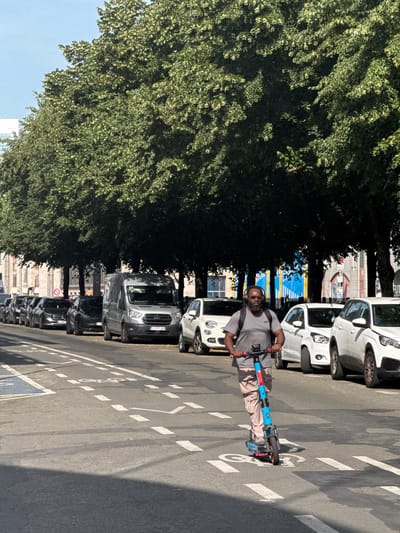 A man is riding an electric scooter down a street in Brussels, Belgium. He is wearing a grey t-shirt, light-colored shorts, and glasses. He has a backpack on. The scooter is blue and red. The man is in the middle of the street, which is paved with asphalt and marked with white lines. There are cars parked on the side of the street, and a line of trees with green leaves casting shadows on the road. The sky is blue, suggesting it is daytime and the weather is sunny. Buildings are visible in the background. The overall scene appears to be a typical urban environment on a pleasant day. The license plate on the grey van visible in the image reads "1 YTS 460".