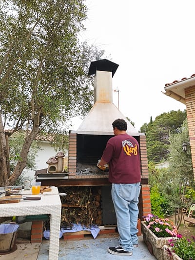 A man in a burgundy t-shirt and blue jeans stands at a brick grill in a backyard setting in Sant Esteve Sesrovires, Spain. He appears to be tending to the grill, holding a container. The grill has a metal chimney and contains wood and charcoal. A white table with items like food, utensils, and a glass of juice sits to the left of the grill. There are trees and greenery surrounding the grill area, and part of a house is visible to the right. A planter filled with flowers is located near the man. A decorative mushroom house is on the grill's countertop.