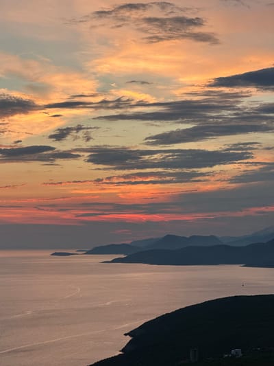 The image depicts a coastal landscape at sunset, with a focus on the sky and the sea. The sky is filled with dramatic clouds painted in hues of orange, pink, and gray. The sun, though not visible, casts a warm glow across the scene. Below the sky, the calm sea reflects the colors of the sunset, with subtle ripples and patterns on its surface. Silhouetted mountains form a dark, undulating line across the horizon, separating the sea from the sky. In the foreground, a dark, lush hillside slopes down towards the water, with a few small structures barely visible at its base. The overall atmosphere is serene and picturesque, characteristic of a coastal sunset in a Mediterranean-like setting. The location is identified as Kovačko Polje, Montenegro.