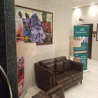 In the interior of what appears to be the Jogor Hotel in Ibadan, Nigeria, a dark brown leather sectional sofa with four cushions occupies the foreground. To the left, a standing lamp with a cream-colored cylindrical lampshade is positioned in front of a large, framed painting depicting three figures in traditional attire. The painting's background is a vibrant swirl of colors. To the right of the sofa, a green banner stands upright, featuring the Jogor Hotel logo and information about its facilities, including "LUXURY ROOMS | RESTAURANT | GYM | POOL | BAR | SALON & SPA | CHILDREN PLAY ROOM". A phone number, "09159999400," and details about "BUFFET BRUNCH EVERY SUNDAY" are also visible. Adjacent to the banner, a sign with male and female restroom symbols and a wheelchair accessibility symbol is mounted on the wall. The floor is tiled with light-colored square tiles. The walls are paneled with off-white tiles.
