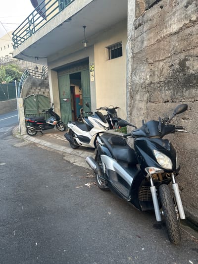 The image depicts a street scene in Câmara de Lobos, Portugal, featuring a row of parked scooters. Three scooters are visible: one black, one white, and one dark-colored scooter parked behind the others. They are parked on the side of the road next to a building with a concrete exterior. A barred window is visible on the building. The scooters are parked on the asphalt road. The lighting appears to be natural daylight.