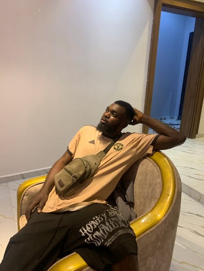 An adult male is depicted seated in an indoor setting. The specific location of Dong, Nigeria, cannot be independently confirmed from the image. The subject has dark skin, short dark hair, and a beard. He is wearing a light peach-colored t-shirt with an Adidas logo and a Manchester United crest, black shorts with white text, and a green canvas crossbody bag worn over his chest. He is seated in a plush armchair featuring brown/beige upholstery and prominent gold-colored armrests. The man is leaning back, with his left arm bent and hand near the back of his head, looking upwards and to his left. The room has light-colored walls and a white tiled floor with a grey/black marble-like pattern. A partially open wooden doorway is visible in the background, revealing a dimly lit interior with a blue hue.