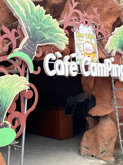 This is a photograph of the entrance to a cafe and camping establishment in Hoa Lư Ward, Vietnam. The entrance features a large, rustic, reddish-brown, artificial rock formation. A prominent sign above the entrance reads "Cafe Camping" in large, white, 3D lettering. To the left and right of the sign are decorative elements resembling palm fronds, rendered in green and yellow. Between the palm fronds and the "Cafe Camping" sign, a smaller sign depicts a cartoon drawing of a child eating, with Vietnamese text that appears to read "An Vặt Ngon Quen Tôi" (Delicious Snacks I'm Familiar With). A silver ladder leans against the right side of the rock formation. In the background, a dark opening suggests the interior of the establishment, with a glimpse of what looks like a wooden counter. The ground in front of the entrance is paved.