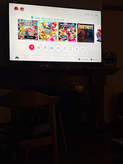 The image shows a Nintendo Switch screen displaying the home menu. The screen shows icons for games such as Fortnite, Mario Kart 8 Deluxe, Super Mario Party Jamboree, and Super Smash Bros Ultimate, which is currently running ("En uso" is displayed next to it). The UI is in Spanish ("Opciones," "Iniciar"). The time on the device is 20:07.

The Nintendo Switch is sitting on a table in a dim room. It is not possible to confirm the location in the city of Naucalpan de Juárez, Mexico based on the visual evidence. Other objects in the background, such as a wooden chair and other furniture, are only partially visible due to the poor lighting.