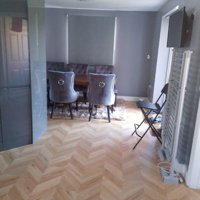 The image shows a residential interior, likely a dining room, in Corby, United Kingdom. The room is decorated in shades of gray and light wood. A dining table is surrounded by upholstered chairs with studded trim and ring pulls. A gray area rug sits beneath the table. The floor is light wood in a herringbone pattern. In the foreground, there is a gray kitchen unit to the left. To the right is a folding chair and a white electric radiator. A flat-screen TV is mounted on the wall. The blinds are closed. There are no people visible in the shot.
