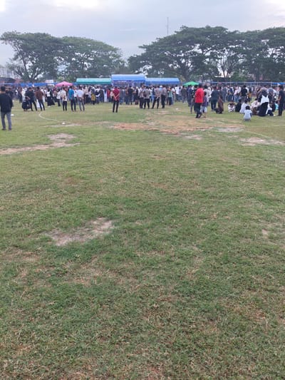 The image shows a large, outdoor gathering of people in a grassy field in Desa Meunasah Blang, Indonesia. The ground is covered in grass with patches of dry, bare earth. In the background, there are several tents set up, some of which appear to be advertising or for vendors. A few trees with full canopies are visible behind the tents. The sky is overcast, suggesting it may be late afternoon or evening.
A crowd of people, estimated to be over a hundred, are spread across the field. Many are standing and appear to be engaged in conversation or watching something. Some individuals are seated on the grass. The demographic appears mixed, with men, women, and children present. Clothing styles are varied, with some people wearing traditional Indonesian attire and others in casual Western clothing. Several individuals appear to be law enforcement officers, identifiable by their uniforms.
The overall scene suggests a public event, possibly a fair, festival, or community gathering. The presence of tents and the concentration of people indicate some form of organized activity is taking place within the field. The ground shows signs of wear, suggesting it is a frequently used public space.