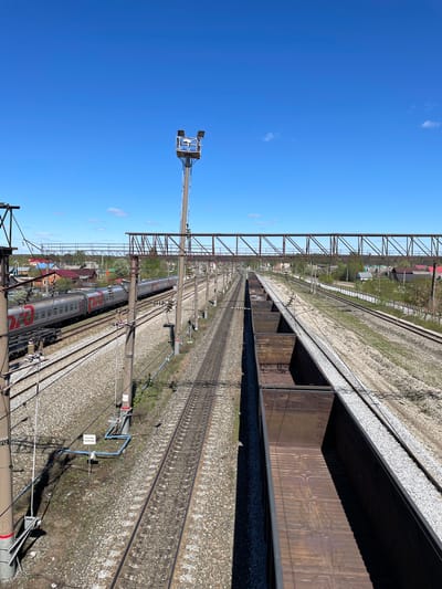 The image shows a railway yard in Kizner, Russia, on a clear day. Several parallel tracks extend into the distance, covered in gravel. A line of open-topped freight cars sits on one track, their rusted metal interiors visible. Overhead power lines and supporting structures run alongside the tracks. In the distance, there's a passenger train, identifiable by its silver cars with red stripes. A tall metal tower stands to the left, equipped with lighting fixtures. Buildings and greenery are visible in the background.
