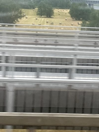 The image shows a blurry view of a field with hay bales, seen from what appears to be a moving vehicle, possibly a train or car, on a bridge. The field is golden yellow with scattered round bales of hay. Trees and vegetation are visible in the background, and a bridge structure is seen in the far distance, suggesting an urban or semi-urban setting. The foreground consists of multiple horizontal metallic structures, likely parts of the bridge railing or the vehicle's interior. The overall impression is one of travel and a fleeting glimpse of the countryside. This scene is consistent with areas near the Elbe River in Magdeburg, Germany, which has many bridges and green spaces suitable for agriculture.