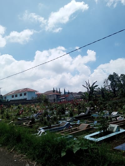 The image depicts a cemetery located in Kioumaknao 2, Laos, under a sky with scattered cumulus clouds. In the foreground, numerous graves are visible, some framed with light blue borders. Lush greenery, including grass and various plants, surrounds the graves. In the background, several buildings are present, including a light blue structure with a red roof. An electrical wire stretches across the upper portion of the frame. The scene is bathed in daylight.