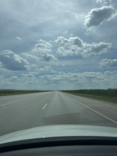 The image captures a straight, asphalt road with white lane markings, likely a highway, extending into the distance. The perspective suggests the photo was taken from inside a vehicle, as the hood of a white car is visible in the foreground. To the left of the road, a field of sunflowers stretches towards the horizon. To the right, a dense green crop, possibly corn, lines the roadside. The sky is filled with a dramatic array of cumulus clouds against a pale blue backdrop, hinting at a partly cloudy day. The background shows a distant, hazy landscape of low hills or mountains. The road appears to be in a rural area outside the city of Byala Slatina, Bulgaria. No people are clearly visible on the road or in the surrounding fields.