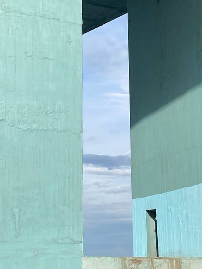 An abstract architectural photograph captures a segment of a light blue sky with white clouds, framed horizontally by concrete structures. The dominant color of the concrete elements is a muted greenish-blue or teal. The upper and lower sections are formed by broad concrete slabs, likely part of a bridge or underpass.

On the upper slab, a diagonal shadow casts a darker tone across its surface, indicating strong, directional sunlight. The concrete shows visible texture from its construction or weathering, with subtle variations in its painted finish. To the far right, a vertical concrete element, possibly a pillar or wall, appears in a lighter, more natural beige-brown tone, contrasting with the greenish-blue. A small, dark, rectangular cutout or opening is present in the upper right concrete structure.

The central horizontal band reveals a bright daytime sky, suggesting sunny weather with scattered cumulus clouds. There are no people, visible activities, or discernible text in the image. The scene is set in Grigor'evskoe, Russia, under bright daylight conditions.