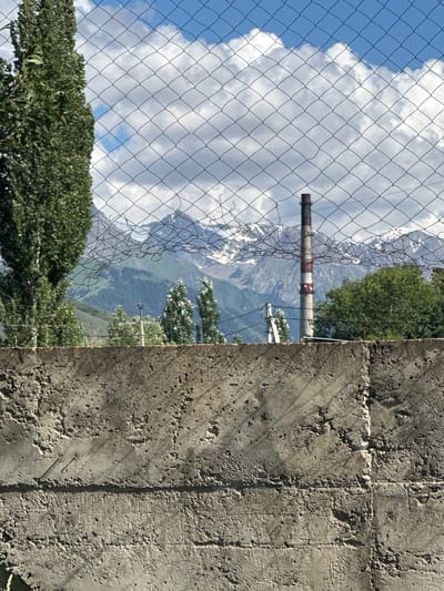 This image depicts a view through a chain-link fence towards a mountainous landscape. In the foreground, a rough, textured concrete wall occupies the right side of the frame. To the left, the chain-link fence is visible, with lush green trees at the bottom. Beyond the fence, a towering, snow-capped mountain range dominates the background, partially obscured by clouds. A red and white striped chimney, likely an industrial remnant, stands prominently in the middle ground, silhouetted against the mountains. The sky is a vibrant blue, dotted with fluffy white clouds, suggesting a clear and bright day. The overall scene evokes a sense of natural grandeur juxtaposed with signs of human presence or industrial activity. The lighting indicates it is daytime, likely late morning or early afternoon, given the strong shadows cast by the fence and the sunlight illuminating the concrete. The location context of Karakol City, Kyrgyzstan, suggests this might be a view from the outskirts of the city, where urban infrastructure meets the rugged natural beauty of the Tian Shan mountains. There is no visible text in the image.