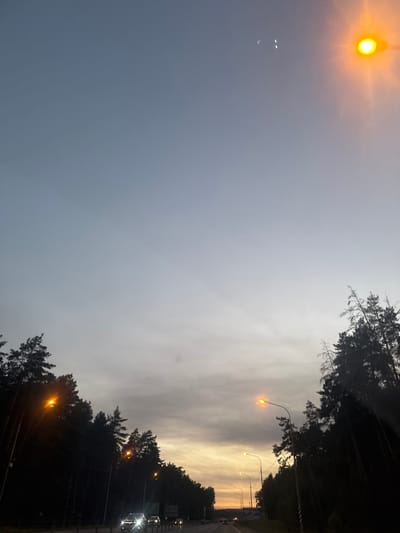 The image shows a road at dusk, lined with dark trees on both sides. Several streetlights illuminate the scene with an orange glow. Cars are visible on the road, with their headlights on. The sky is a gradient of grey and pale orange, with some clouds. The location is identified as Mstikhino, Russia.