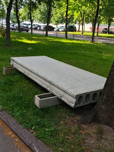 A large, hollow concrete slab is resting on two wooden blocks on a grassy area. The slab is angled, with one end closer to the viewer and the other end further away. A tree trunk is visible to the right of the slab. In the background, a row of cars is parked along a street, separated from the grassy area by a metal fence. Trees line the street, and some buildings are visible in the distance. The scene appears to be outdoors during the daytime, with sunlight casting shadows on the grass. The ground is covered in green grass with some small white flowers. The concrete slab is a rough, light gray color and has a series of circular hollows running along its length. The wooden blocks appear weathered. The overall setting seems to be a residential or public area with a park-like feel.