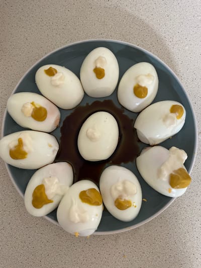 An overhead shot shows a dark gray plate filled with eleven hard-boiled eggs, each topped with a dollop of mayonnaise and a smaller dollop of mustard. The eggs are arranged in a circular pattern, with a dark liquid, possibly soy sauce, pooled in the center of the plate. The plate rests on a light-colored, speckled countertop.