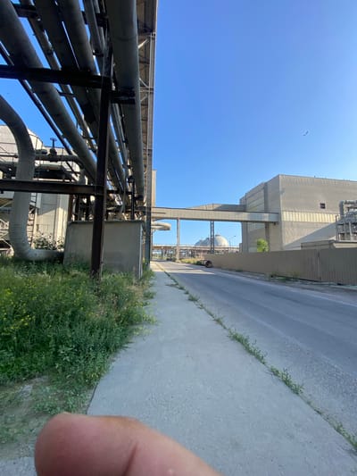 The image depicts an industrial area with a clear blue sky. In the foreground, a concrete sidewalk runs parallel to a road. To the left of the sidewalk, dense green vegetation with small yellow flowers grows. Above the vegetation and sidewalk, a complex network of large, grey pipes supported by metal structures is visible, running horizontally and angled upwards.

Further into the scene, a long, grey, elevated enclosed walkway connects two industrial buildings. The building on the right is a large, rectangular structure with a textured facade and bands of windows. A metal fence runs along the edge of the property. In the distance, two large, domed structures are visible, characteristic of industrial or storage facilities. A single bird is captured in flight in the sky. The overall impression is an industrial landscape under a bright sky.

This location is identified as the Police Industrial Zone in Police, Poland.