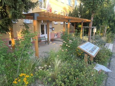 The image depicts the exterior of a building in Galanta, Slovakia, featuring a modern wooden pergola extending from the entrance. The building has a yellowish facade with multiple windows and flags displayed above the entrance. In front of the building, there is a landscaped area with various plants, including flowers, grasses, and shrubs. A metal bench sits on a paved walkway beneath the pergola. A wooden post with an informational sign is positioned in the landscaped area, displaying text and images. The overall scene suggests an outdoor space that is well-maintained and potentially serves a public function.