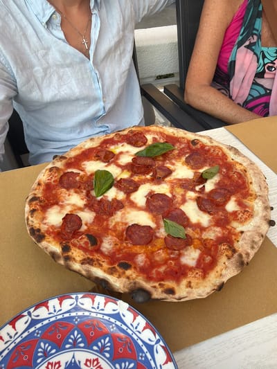 The image captures a close-up of a pepperoni pizza, presented on a light brown surface. The pizza has a golden-brown crust with some charred spots, topped with tomato sauce, melted cheese, and circular slices of pepperoni. A few fresh green basil leaves are scattered across the pizza. In the background, partially visible, is a person wearing a light blue, unbuttoned collared shirt with a silver cross necklace. To the right, another person is visible wearing a brightly colored garment, possibly a dress or scarf, with a patterned design. A portion of a decorative plate with red, blue, and white patterns is in the lower left corner of the image. The setting appears to be an outdoor dining area, possibly in Lugano, Switzerland, suggested by the presence of a table and chairs.