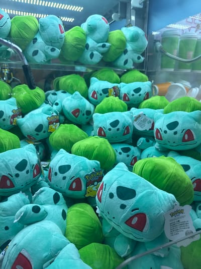 This media file shows a close-up view of a claw machine filled with many plush toys. The subjects are primarily the plush toys, which are predominantly light green Bulbasaur characters from the Pokémon franchise. There are also some larger, lime green round objects, possibly also plush toys or filler material within the machine.
The scene is the interior of a claw machine, indicated by the transparent glass front and the visible claw mechanism in the lower left of the image. The setting is an indoor arcade or entertainment venue, suggested by the bright, artificial lighting, the presence of a claw machine, and the stacked nature of the plush toys within the confined space.
No specific activity or event is taking place, other than the display of the toys within the machine, awaiting selection by a player.
Notable details include the repeated pattern of Bulbasaur plush toys, some of which have red eyes and red lips. Several tags are visible on the plush toys. One tag, in the upper right quadrant, has Chinese characters and what appears to be Pokémon branding. Another tag, in the center left, also displays Pokémon branding. The image is well-lit, suggesting it was taken during operational hours, likely during the day or evening in an indoor environment. The location is specified as Shanghai, China.
Visible text includes "Pokémon" on the tags, along with Chinese characters on at least two tags. Some of the characters on the tag in the upper right appear to be manufacturer information and possibly a product code. There is also some text on a label on the right side of the machine, but it is too blurry to read.