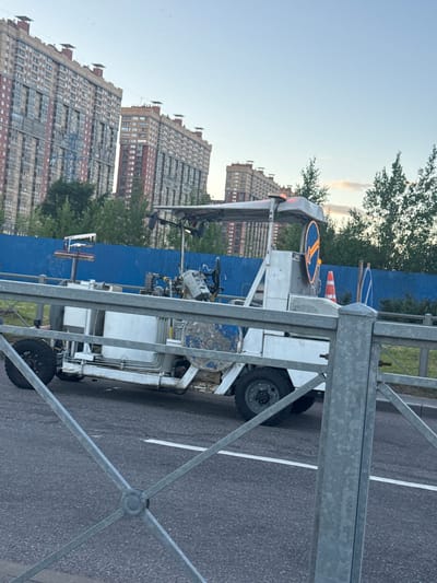 In Pargolovo, Russia, an abandoned white line marking machine sits on a paved road. The machine is equipped with a canopy, a large tank, and a rotating arrow sign with a light border. Construction fencing and trees line the background, with tall apartment buildings visible in the distance. The sky is clear and blue.